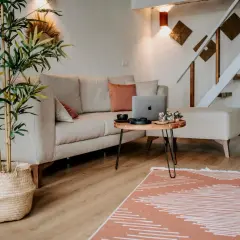 Przytulne, małe mieszkanie w stylu boho. Jasna kanapa, drewniany stolik z laptopem i ozdobami, bambus w koszu, schody i rośliny tworzą ciepłą atmosferę.