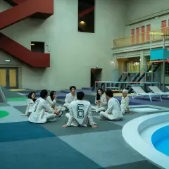 Grupa ludzi w białych strojach z numerami siedzi na podłodze w surrealistycznym otoczeniu, przypominającym scenografię z "the 8 show".