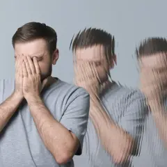 Mężczyzna zakrywa twarz dłońmi, jego postać rozpada się, sugerując stan delirium.