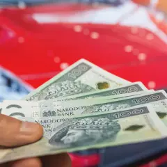 Dłoń trzyma banknoty 100 zł na tle czerwonego samochodu. Koszty posiadania auta, w tym podatek od samochodu, mogą być znaczące.