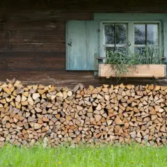 Ein ordentlicher Stapel Holz liegt vor der rustikalen Hauswand. Ein Fenster mit blauen Fensterläden und einem Blumenkasten mit Kräutern darüber.
