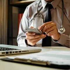 Lekarz w koszuli i krawacie, ze stetoskopem na szyi, korzysta z telefonu. Może szuka informacji, jak napisać opinie o lekarzu, lub sprawdza kalendarz wizyt.