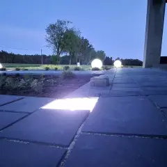 Nocny ogród rozświetlony kulistymi lampami. Ścieżka z kostki brukowej, na której widać wbudowane ledy, prowadzi do domu.