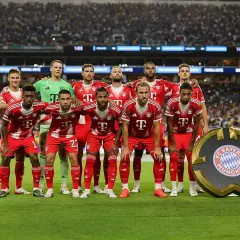 Drużyna Bayern Monachium – zawodnicy w czerwonych strojach pozują do zdjęcia na tle trybun. Obok nich stoi okrągłe logo klubu.