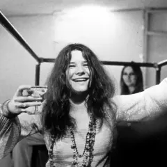 Janis Joplin, z kieliszkiem w dłoni, uśmiecha się szeroko. Jej charakterystyczne włosy opadają na ramiona, a na szyi wisi naszyjnik. W tle widać inną kobietę. To zdjęcie przypomina o jej niezapomnianych **janis joplin utworach**.