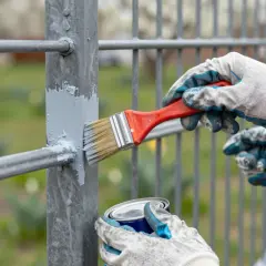 Ręce w rękawiczkach malują pędzlem stare ogrodzenie na szaro. W tle zielony ogród z żółtymi kwiatami.