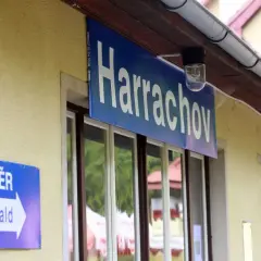Tabliczka "Harrachov" nad oknami dworca. Obok niebieski znak "SMĚR Tanvald" z białą strzałką. Pociąg Szklarska Poręba Harrachov czeka na pasażerów.
