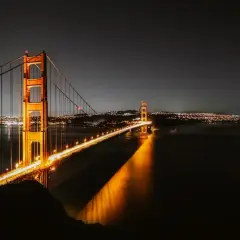 Złota Brama nocą, oświetlona, nad wodą. W oddali widać miasto w Kalifornii, San Francisco, z migoczącymi światłami.