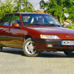 Czerwony Daewoo Espero z lat 90. na tle zieleni i brukowanej drogi.