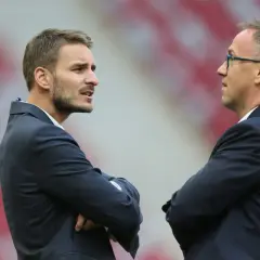 Dwaj mężczyźni w garniturach rozmawiają na tle stadionu. Jeden z nich, z zarostem, to dyrektor sportowy.