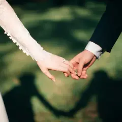 Dłonie nowożeńców splecione w uścisku, symbolizującym przysięgę małżeńską. Na palcu panny młodej lśni pierścionek.