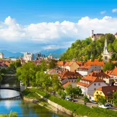 Widok na rzekę Ljubljanicę, mosty i zabytkowe budynki Lublany ze wzgórzem zamkowym w tle.