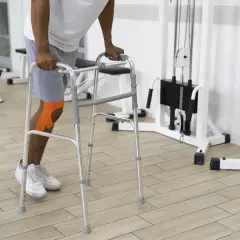 Mężczyzna z pomarańczową taśmą na kolanie ćwiczy z chodzikiem. Różne rodzaje sprzętu ortopedycznego pomagają w rehabilitacji.