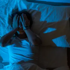 Persona en cama, con las manos en la cara, iluminada por luz azul. Las sombras en la pared sugieren que es por eso que me cuesta dormir.