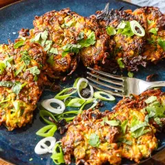 Knusprige zucchinipuffer mit kartoffeln, garniert mit frischen Kräutern und Frühlingszwiebeln, liegen auf einem blauen Teller. Eine Gabel liegt daneben.