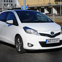 Białe małe auto, Toyota Yaris, jedzie po drodze. W tle budynki i torowisko tramwajowe.