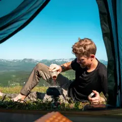 Man häller kaffe i en kastrull vid tältöppningen. En del av packlista camping. Vacker utsikt över fjällen.