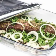 Mięso w naczyniu żaroodpornym, przykryte folią aluminiową, która pomaga utrzymać ciepło. Cebula i zioła dodają smaku.