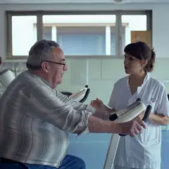 Une aide-soignante encourage un homme âgé sur un vélo d'appartement. Une scène qui pourrait figurer dans une lettre de motivation pour travailler auprès des personnes âgées.