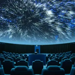 Wnętrze planetarium, gdzie na kopule wyświetlane są rozgwieżdżone niebo i galaktyki. Widownia z niebieskimi fotelami czeka na seans.