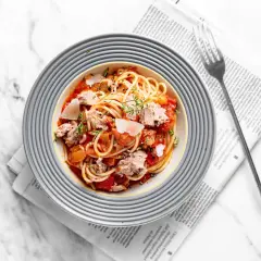 Pyszny makaron z tuńczykiem i pomidorami, posypany parmezanem i ziołami, podany na talerzu na tle gazety.