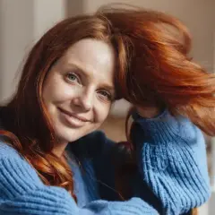 Uśmiechnięta rudowłosa kobieta w niebieskim swetrze, jej rude włosy falują. Inspirujące rude włosy stylizacje.