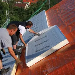 Dwaj pracownicy montują płyty izolacyjne na dachu. To kluczowy etap prac nad ociepleniem nakrokwiowym, zapewniającym komfort termiczny budynku.