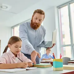 Nauczyciel z brodą, trzymający laptopa, pomaga uczennicy w pisaniu. W tle inni uczniowie. To jego legitymacja nauczyciela.