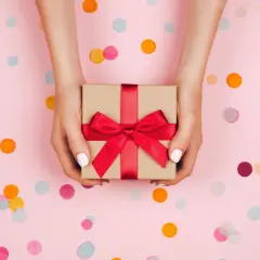 Dłonie trzymające prezent z czerwoną wstążką. Tło w kolorze różowym z kolorowym konfetti. Idealne na religijne życzenia urodzinowe.