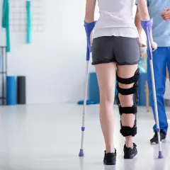 Pacjentka z ortezą na nodze i kulą ortopedyczną, ucząca się chodzić. Jakie kule ortopedyczne wybrać? Fizjoterapeuta pomaga w rehabilitacji.
