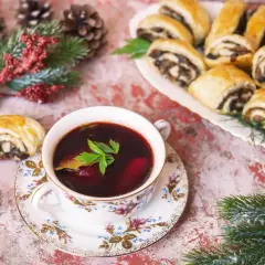 Aromaticzny barszczyk czerwony z pasztecikami, ozdobiony gałązką zieleni, czeka na świątecznym stole wśród szyszek i gałązek.