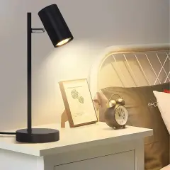 Lampara mesilla negra ilumina un cuadro y un despertador vintage en una mesita de noche junto a una cama.