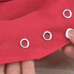 Demonstration of how to put metal rings in fabric. A hand holds red fabric with several silver grommets already attached, and another grommet is about to be placed.