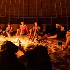 Grupa ludzi siedzi na piasku w okrągłej, drewnianej chacie, otoczona świecami. Rozmawiają i śmieją się, przygotowując się do ceremonii. Ayahuasca cena może być różna, ale to doświadczenie jest bezcenne.