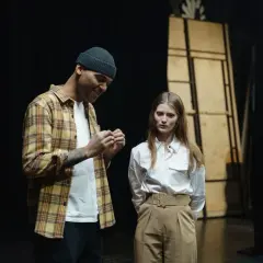 Mężczyzna w kratę i czapce rozmawia z kobietą na scenie. Czy to próba przed występem?