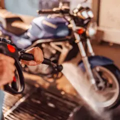 Persona limpiando una moto azul con una hidrolavadora. El agua salpica sobre la rueda delantera.