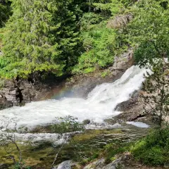 Vattenfall i Idre, med en regnbåge som skimrar i dimman från det forsande vattnet. Omges av grönskande träd och klippor.
