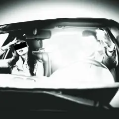Frau im Auto, die sich die Hand ans Gesicht hält, während ein Mann am Steuer sitzt. Ein grelles Licht, wie von einem Blitzer, überstrahlt die Szene.