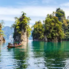Bootfahrt durch smaragdgrünes Wasser im **Thailand Regenwald**, vorbei an steilen, bewachsenen Felsen unter blauem Himmel.