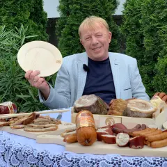 Mirek Szołtysek prezentuje bogactwo wędlin i mięs na drewnianych deskach, trzymając w dłoni pusty talerz.