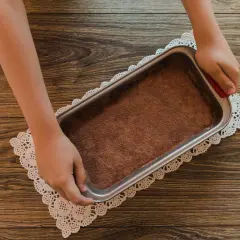 Mani che preparano un tiramisù senza caffè, con cacao in polvere in una teglia su un centrino di pizzo.