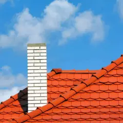 Pomarańczowy dach wielospadowy z ceglanym kominem na tle błękitnego nieba z chmurami.