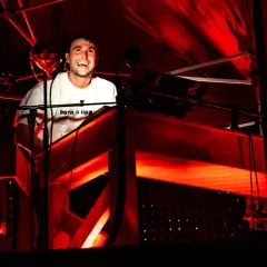 Il **best techno dj** ride dietro la console, immerso in luci rosse intense.