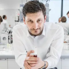 Mężczyzna w białej koszuli trzyma fiolkę z płynem, w tle laboratorium, gdzie pracują inni naukowcy. To kluczowe xtpl akcje.