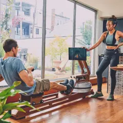 Para ćwiczy na ergometrze wioślarskim, który wygląda jak rower dla bliźniaków. Kobieta stoi obok, patrząc na ekran.