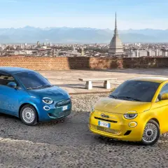 Dwa Fiaty 500 hybryda, niebieski i żółty, stoją na brukowanym placu z widokiem na panoramę miasta i góry.
