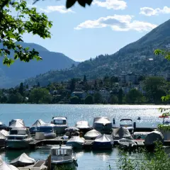 Båtar täckta med presenningar vid en brygga vid Lago Maggiore. Berg och en stad syns i bakgrunden.