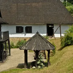 Skansen w Dobczycach: drewniane chaty, studnia i zielone wzgórza tworzą sielski krajobraz.