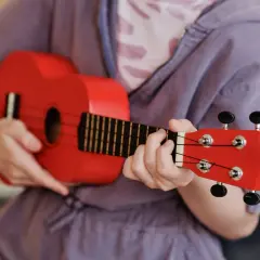 Enfant jouant d'un petit instrument de musique rouge vif, un ukulélé, avec concentration.