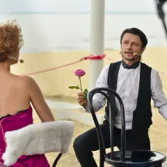 Aktor z Teatru Czwarte Miasto trzyma różę, zwracając się do widowni. Kobieta w fuksjowej sukni siedzi tyłem.